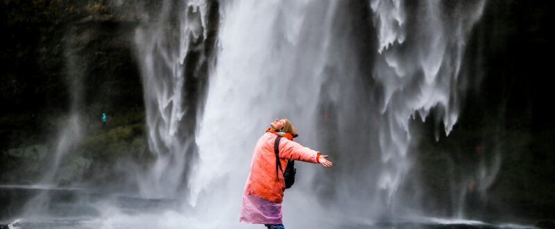A traveler stands under a waterfall with arms outstretched, enjoying the unique guest experience offered as an extra with their accommodation booking.